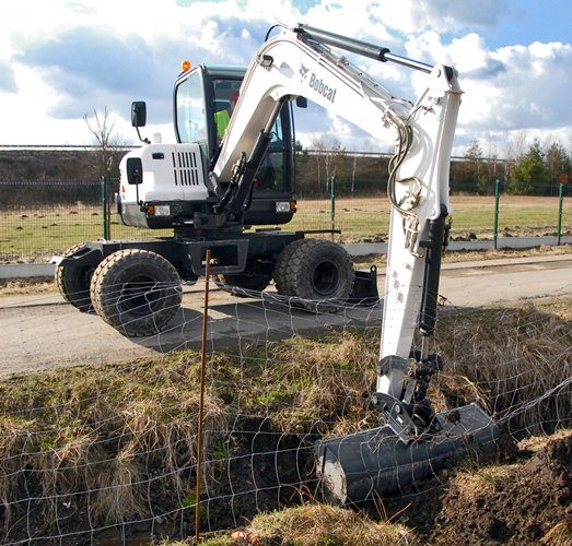 White wheeled excavator working beside a roadside fence