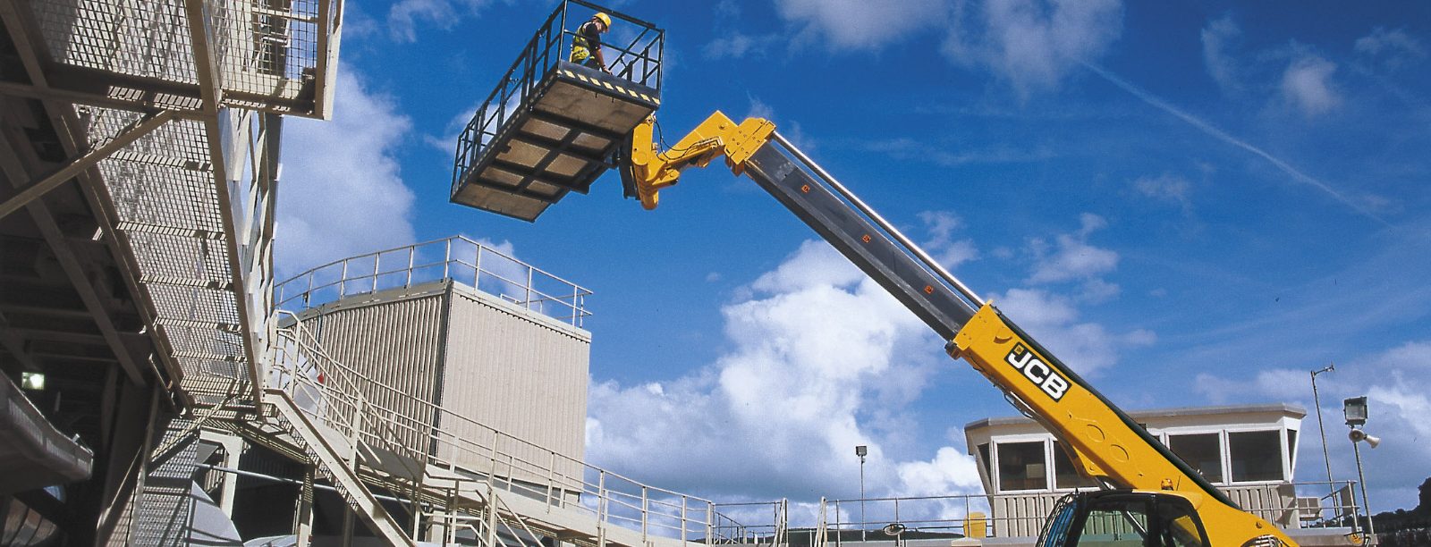 JCB telehandler lifting a work platform beside an industrial structure